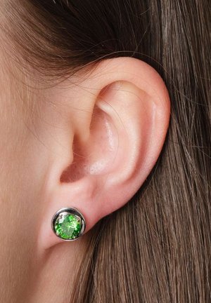 Nahaufnahme eines rechten Ohrs mit glattem braunem Haar, das einen runden silbernen Stecker mit einem facettierten hellgrünen Edelstein trägt.