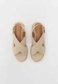 Beige espadril dengan tali silang yang memiliki tampilan suede, dilengkapi dengan sol jute dan aksen logam kecil pada tali. Desain datar.