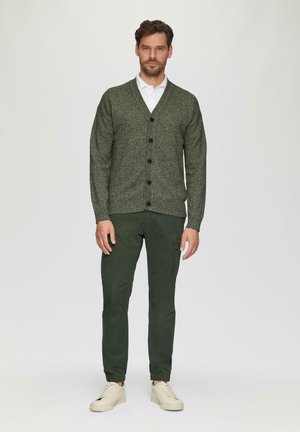 Strukturierte grüne Strickjacke mit sechs schwarzen Knöpfen, kombiniert mit grünen Hosen und weißen Turnschuhen, getragen von einem Mann, der vor einem neutralen Hintergrund steht.