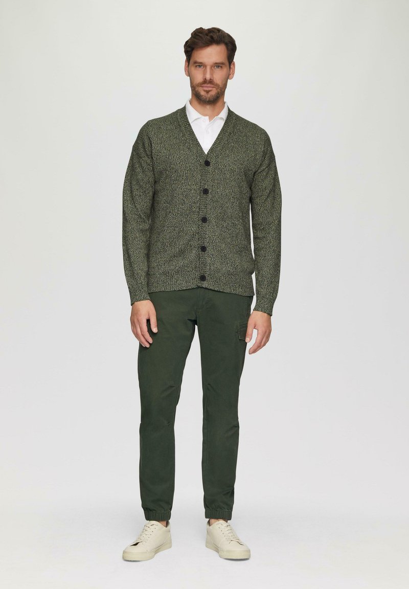 Strukturierte grüne Strickjacke mit sechs schwarzen Knöpfen, kombiniert mit grünen Hosen und weißen Turnschuhen, getragen von einem Mann, der vor einem neutralen Hintergrund steht.