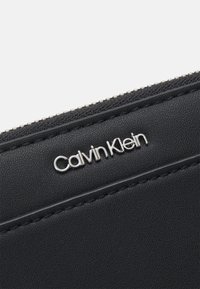 Czarny portfel ze strukturalnej skóry, z srebrnym logo "Calvin Klein", z zamknięciem na zamek i kontrastowym szyciem wzdłuż krawędzi.