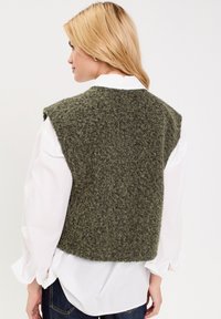 Gilet sans manches en tricot vert olive avec un tissu texturé, porté par-dessus une chemise blanche à col. Détail de franges subtiles le long de l'ourlet.