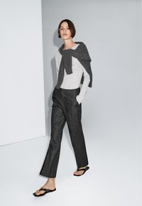 Grijze longsleeve top en donkergrijze broek, met een bijpassende trui over de schouders gedrapeerd. Zwarte slippers maken de outfit compleet.