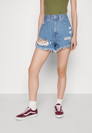 Personne portant un short en jean déchiré taille haute, des chaussettes blanches et des baskets bordeaux, debout devant un fond clair uni.