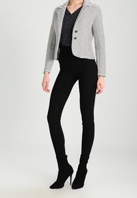 Pruhovaný blazer v bílé a černé látce se dvěma knoflíky, doplněný tmavým třpytivým topem a černými upnutými kalhotami, končícími na jehlových podpatcích.
