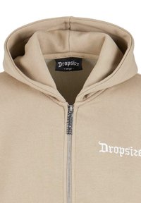 Beige hoodie med lynlås foran, blød tekstur, stor hætte og broderet "Dropsize" logo i hvid. Sort etiket indeni.