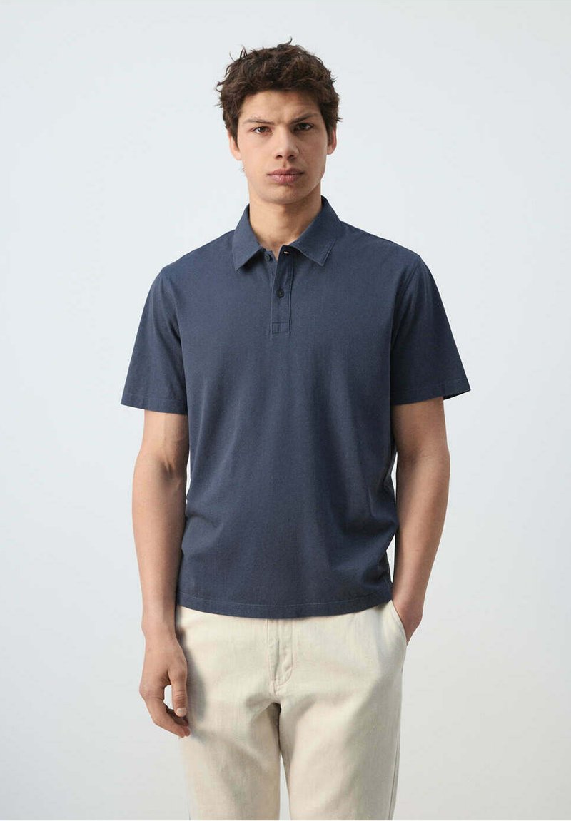 Jeune homme aux cheveux courts et foncés portant un polo bleu foncé à manches courtes et un pantalon beige clair, debout avec une main dans la poche.
