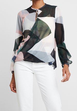 Donna che indossa una camicetta trasparente a maniche lunghe con motivi geometrici astratti in nero, bianco, verde e rosa abbinata a pantaloni bianchi.