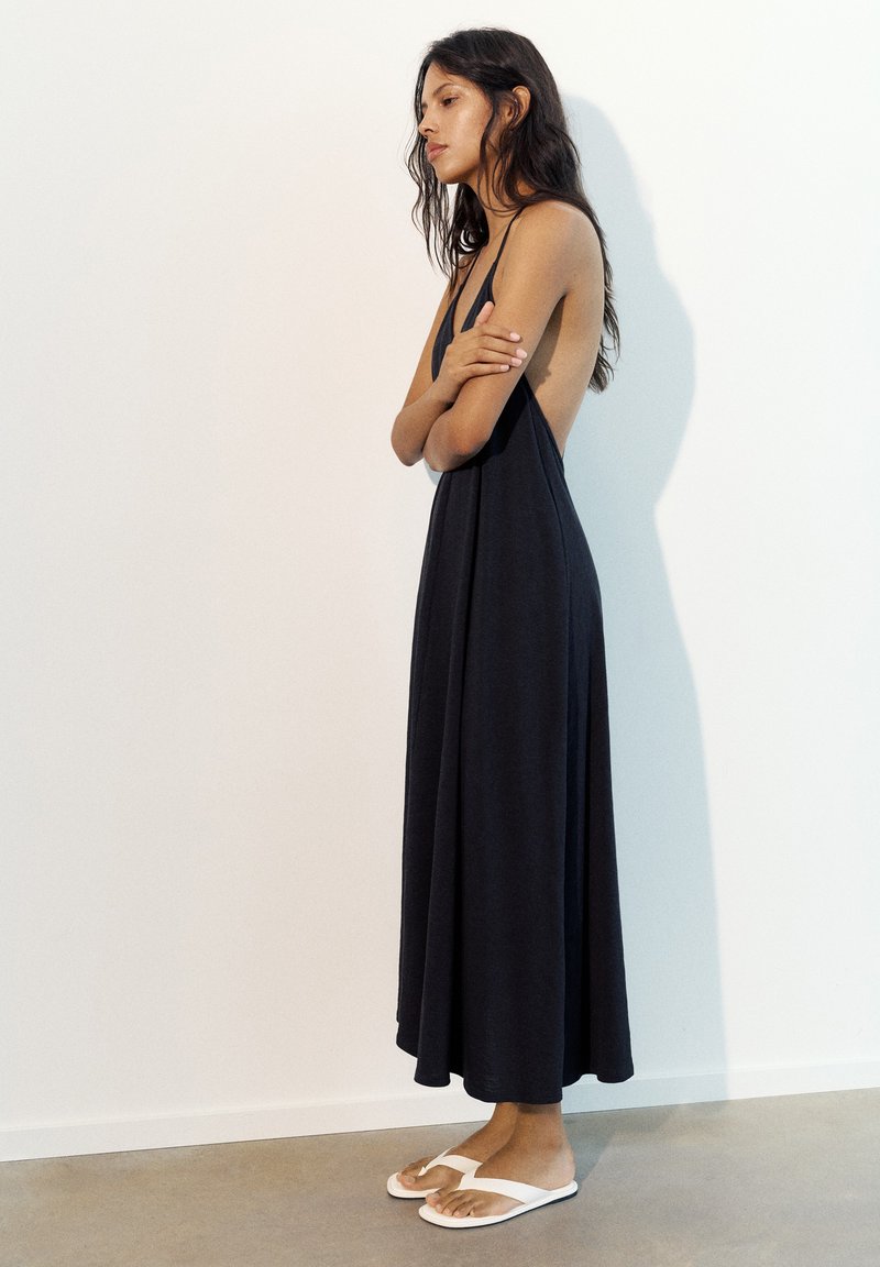 Femme debout de profil portant une robe maxi noire sans manches et des sandales blanches devant un mur clair uni.