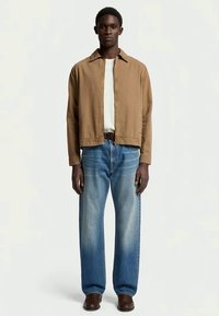 Veste légère marron sur un t-shirt blanc, associée à un jean bleu clair à jambes larges et des chaussures marron. Design simple et épuré.