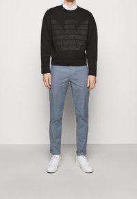 Černá mikina se zvýšeným logem orla, v kombinaci se světle modrými kalhotami a bílými teniskami. Materiál vypadá hladce; střih je ležérní.