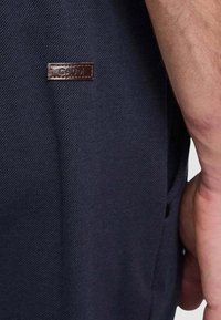 Pantaloni blu navy scuro in tessuto testurizzato con un'etichetta in pelle marrone visibile sul lato con la scritta "GAUDI". Presentano tasche e una vestibilità sartoriale.