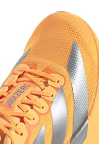 Orange athletic shoe with a textured fabric upper, silver accents, and laces. Features a prominent branding label on the tongue.