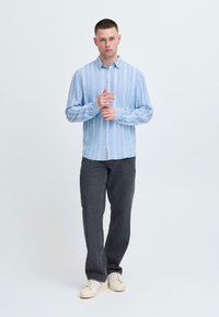 Chemise à rayures bleu clair à boutons avec manches longues, associée à un pantalon gris foncé et des baskets beige, présentée sur un fond uni.