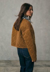 Brun kordjacka i cropped-modell med diamantmönster, med ett kviltat design och kontrasterande detaljer, bärs över en mönstrad topp och blå jeans.