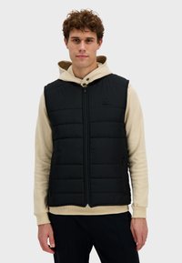 Czarna ocieplana kamizelka z zamkiem z przodu, noszona na jasnobeżowej bluzie z kapturem. Posiada prosty logo i boczne kieszenie.