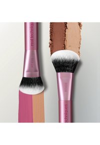Deux pinceaux de maquillage Real Techniques RT459 avec manches roses, l'un tamponnant une crème et une poudre brune, l'autre mélangeant une crème rose et beige.