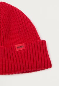 Berretto lavorato a maglia rosso con texture a coste. Presenta un piccolo etichetta rettangolare con la scritta "HUGO" in nero sul risvolto.