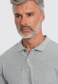 Jasnoszara koszulka polo z kołnierzykiem, z dwu guzikowym zapięciem, miękką teksturą i krótkimi rękawami, zaprezentowana na górnej części ciała modela.