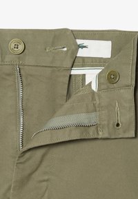 Ceinture de pantalon marron clair avec fermeture éclair partiellement ouverte, deux boutons, et logo Lacoste visible sur l'étiquette intérieure.