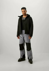 Zwarte jas met capuchon, grijs en zwarte broek met zakken, en zwart schoeisel. Stof lijkt waterdicht met een gestructureerde afwerking.