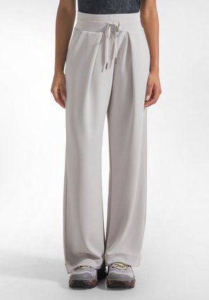 Losse lichtgrijze broek met een trekkoord in de taille, gedragen met grijze en witte sneakers, getoond van taille tot voeten tegen een effen achtergrond.