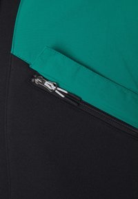 Sezione di abbigliamento nera e turchese con una tasca con cerniera decorata con dettagli in paracord bianco, che offre un contrasto di texture tra i due materiali.
