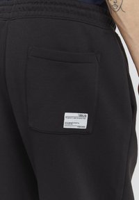 Zwarte joggerbroek met een rugzak met een wit label. Gemaakt van premium stof met een gladde textuur en een elastische tailleband.