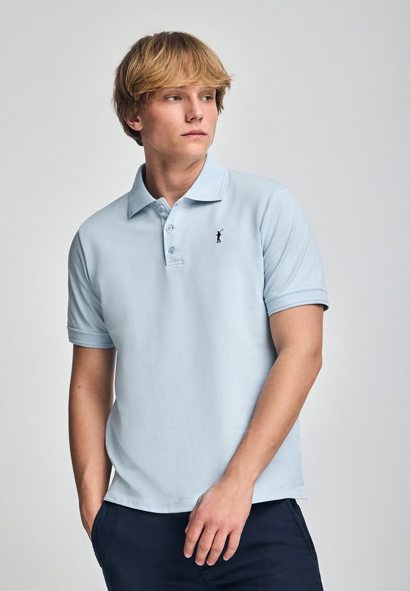 Polo de color azul claro hecho de algodón, con un tapón de tres botones, mangas cortas y un pequeño logo negro en el lado izquierdo del pecho.