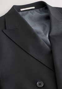 Fekete, szabott blézer éles hajtókával, sima szürke béléssel, apró pöttyökkel, és egyetlen fekete gombbal záródik.