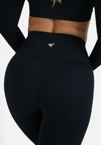 High-waisted black leggings made of textured fabric featuring a subtle logo in white on the back. Designed for a snug fit and flexibility.