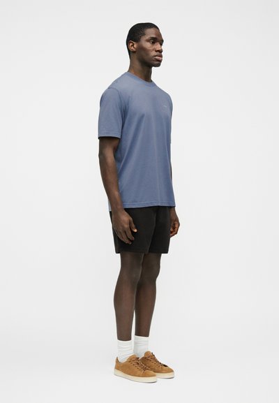 Άνδρας που στέκεται, φορά μπλε μπλούζα, μαύρο σορτς, λευκές κάλτσες και καφέ αθλητικά παπούτσια, μπροστά σε απλό λευκό φόντο.