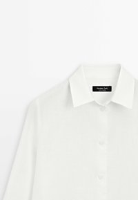 Weißes Leinenhemd mit klassischem Kragen, langen Ärmeln und vorderem Knopfverschluss. Weicher Stoff mit minimalen Nähten. Markenlabel sichtbar.