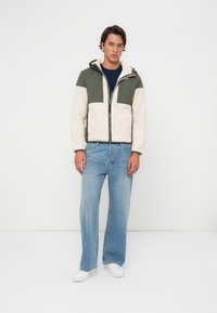 Fleece-fôret jakke med olivengrønne og kremfargede paneler, frontglidelås og justerbar hette, sammen med lyseblå jeans og hvite sneakers.