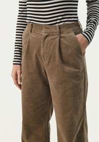 Corduroy-housut lämpimässä ruskeassa sävyssä, joissa on korkea vyötärö, pleat-tied ja sivutaskut, yhdistettynä musta-valkoiseen raidoitettuun toppiin.