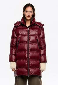 Bordowy puchowy płaszcz z błyszczącym wykończeniem, wyposażony w zamek błyskawiczny, dwie zapinane na zamek boczne kieszenie oraz futrzane akcenty na rękawach.