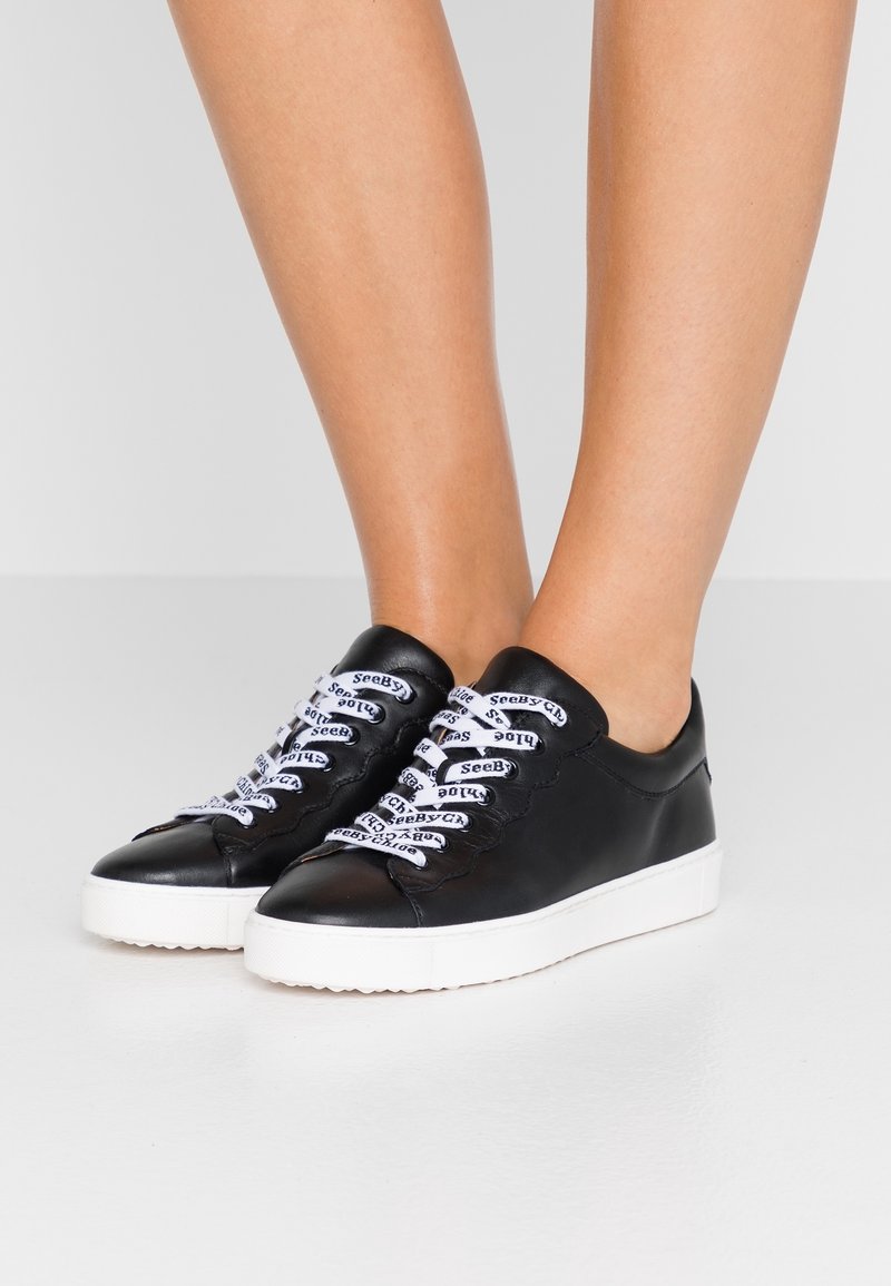 Jambes portant des baskets basses noires avec des semelles épaisses blanches et des lacets blancs imprimés du texte « SeeByChloé » sur fond gris.