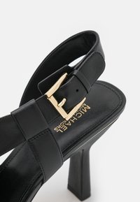 Sandale à talons hauts en cuir noir avec bride à boucle dorée, dos ouvert et logo "Michael Kors" sur la semelle intérieure.