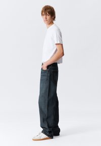 T-shirt branca de manga curta combinada com jeans de ganga escura de corte largo. Os jeans apresentam um design clássico de cinco bolsos e uma lavagem subtil. Ténis brancos.