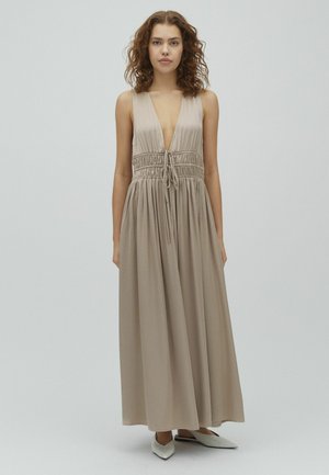 Femme aux cheveux bouclés portant une robe maxi beige sans manches avec un décolleté en V profond et une taille froncée, debout devant un fond clair uni.