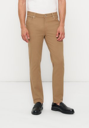 Férfi bézs slim fit nadrágban, fehér ingben és fekete bőr mokaszinban, egyszínű fehér háttér előtt áll.