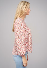 Blusa floreale a maniche lunghe con dettagli arricciati in tonalità di rosa e rosso, realizzata in tessuto leggero, abbinata a jeans azzurri.