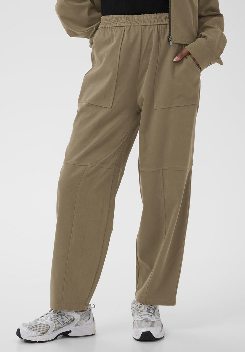 Pantalones cargo caqui con una cinturilla elástica, que cuentan con grandes bolsillos laterales y un corte relajado, combinados con zapatillas blancas.