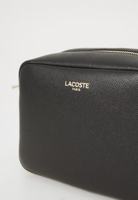 Schwarze, strukturierte Leder-Rechtecktasche mit goldfarbener Reißverschluss und goldenem Lacoste Paris-Logo auf der Vorderseite.