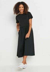 Vestido negro de algodón con mangas cortas, un cuerpo ajustado y una cintura fruncida, con un diseño fluido de pierna ancha. Zapatillas blancas casuales.