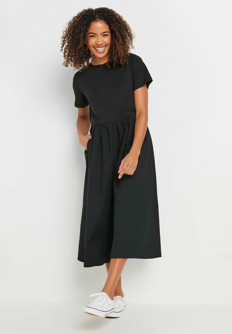 Vestido negro de algodón con mangas cortas, un cuerpo ajustado y una cintura fruncida, con un diseño fluido de pierna ancha. Zapatillas blancas casuales.