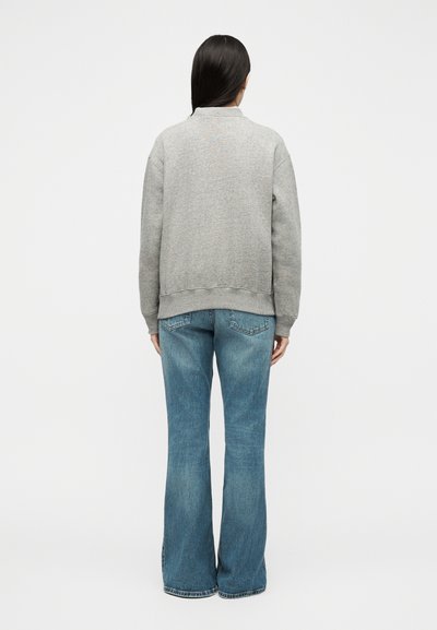 Άτομο με μακριά σκούρα μαλλιά, ντυμένο με γκρι φούτερ και μπλε καμπάνα τζιν, στέκεται γυρισμένο με την πλάτη του σε λευκό απλό φόντο.