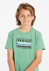 Groene t-shirt van zacht materiaal, met een zwart rechthoekig logo waarop het woord "PROTEST" staat en een meerkleurige golfgrafiek.