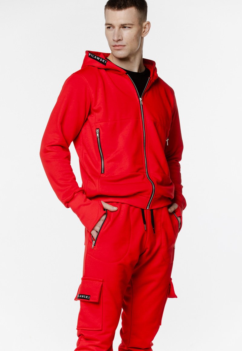 Czerwona bluza zapinana na zamek i dopasowane dresy wykonane z miękkiego materiału. Bluza ma logo na kołnierzu oraz zamykane na zamek kieszenie w obu częściach.