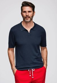 Mörkblå kortärmad polo med krage och v-ringning, bärs med röda shorts med dragsko. Slät textur, avslappnad design.